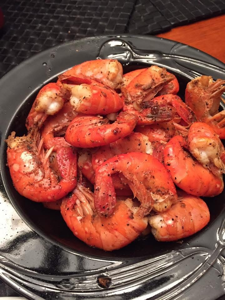 A silver-colored dish containing a cooked catch of prawns, the dish is on a wooden table with black placemats. The prawns are orange in color and are seasoned with flakes of black pepper.